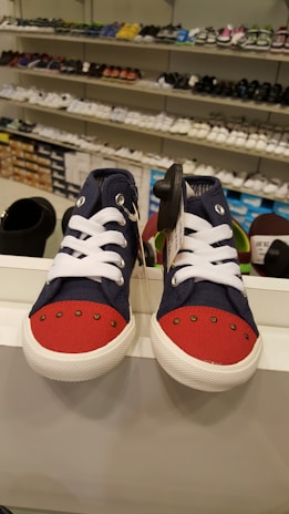 A display features a pair of children's sneakers with navy blue fabric and bright red toes, white laces, and white soles. They are placed on a shelf in a shoe store with other footwear visible in the background, arranged neatly on several shelves.