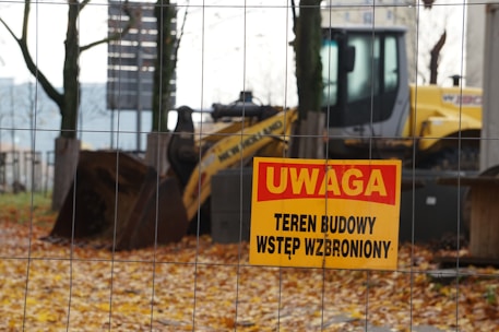 A construction site is behind a wire fence, with a warning sign in Polish that reads 'UWAGA TEREN BUDOWY WSTĘP WZBRONIONY' in the foreground. The background shows a yellow construction vehicle, possibly a backhoe loader, and the ground is covered with fallen autumn leaves.