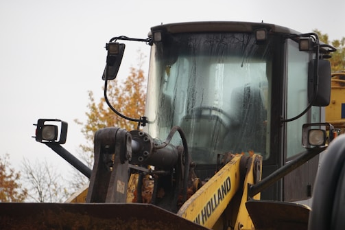 A close-up view of a yellow heavy machinery vehicle, likely a backhoe loader, with visible raindrops on its front windshield. The vehicle has various mechanical parts and equipment attached to it, and the background shows autumn trees with leaves of red, orange, and yellow hues.