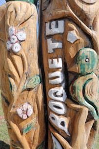 Intricately carved wooden pillar featuring floral and bird designs, with the word 'BOQUETE' prominently displayed in white letters. The wood has a natural finish with visible texture and grain.