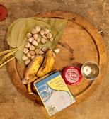 A wooden circular tray displays an assortment of items including two ripe bananas, a cluster of betel nuts on green leaves, a box of sweets with colorful packaging, a small silver cup, and a red container with decorative markings.