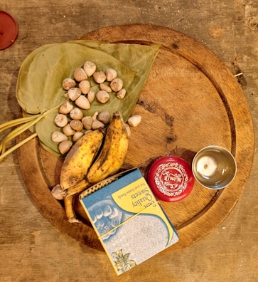 A wooden circular tray displays an assortment of items including two ripe bananas, a cluster of betel nuts on green leaves, a box of sweets with colorful packaging, a small silver cup, and a red container with decorative markings.