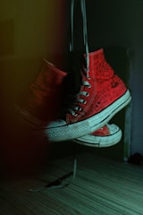 A pair of red high-top sneakers with unique, hand-drawn doodles and writing on them hangs by their laces in a dimly lit room. The lighting creates a dramatic shadow and highlights the textured detail of the canvas material, along with the white rubber soles.