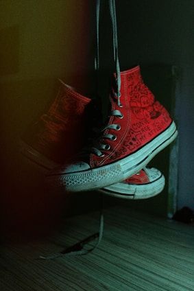 A pair of red high-top sneakers with unique, hand-drawn doodles and writing on them hangs by their laces in a dimly lit room. The lighting creates a dramatic shadow and highlights the textured detail of the canvas material, along with the white rubber soles.