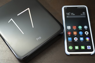 A smartphone with a colorful app-filled screen is placed next to a sleek, black product box labeled '17 Pro'. The surface has a smooth and glossy finish, reflecting light subtly.