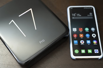 A smartphone with a colorful app-filled screen is placed next to a sleek, black product box labeled '17 Pro'. The surface has a smooth and glossy finish, reflecting light subtly.