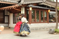 Two individuals are walking along a traditional Korean street, wearing hanbok, which are traditional Korean garments. One wears a vibrant red skirt and a black top, while the other has a blue patterned skirt and a white top. The background features a building with wooden elements and traditional Korean architecture, including tiled roof eaves and paper lanterns. The street is paved with stone tiles.