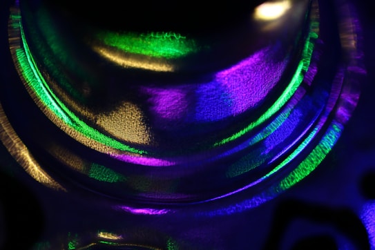 A close-up view of abstract light patterns with vibrant colors creating swirling and overlapping forms. The texture appears smooth and glass-like with reflections and refractions of green, purple, and gold hues.