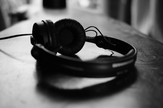 A pair of black over-ear headphones lies on a wooden surface. The headphones are centered in the image, with soft lighting creating a subtle reflection on the table.
