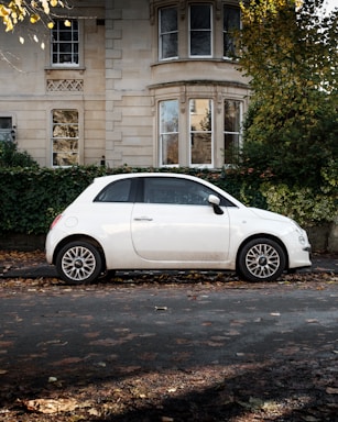 A white compact car is parked on the side of the street in front of a residential building with classic architecture and ivy growing over a low hedge. The ground is covered with fallen leaves, indicating autumn. The building has large windows and stone detailing, giving it an elegant appearance.