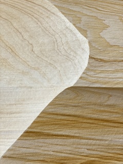 A close-up view of a wooden surface with two overlapping sheets of veneer. The wood grains are distinct and vary in color from light beige to darker brown, creating a natural pattern. The lighting highlights the texture and layers of the veneer.