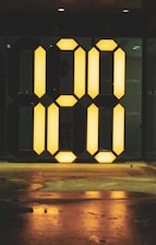 A large illuminated digital-style number display reading '00' is set against a dark background. The glowing yellow numbers are reflected on the wet ground below, creating a contrasting and striking visual effect.