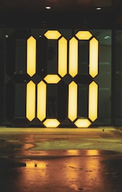 A large illuminated digital-style number display reading '00' is set against a dark background. The glowing yellow numbers are reflected on the wet ground below, creating a contrasting and striking visual effect.