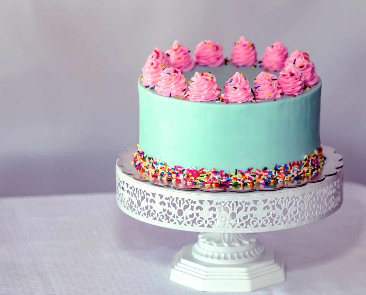 Mint-green cake topped with pink swirls and rainbow sprinkles on a white stand.