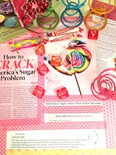 A colorful assortment of candies, including a large rainbow lollipop, Sassy Tarts, Smarties, and Starburst, is displayed on top of a magazine page discussing sugar addiction. Pink, green, and blue plastic spiral toys are placed around the candies. The page contains information about sugar consumption, a quiz on sugar addiction, and a recipe for crack pie.