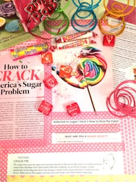 A colorful assortment of candies, including a large rainbow lollipop, Sassy Tarts, Smarties, and Starburst, is displayed on top of a magazine page discussing sugar addiction. Pink, green, and blue plastic spiral toys are placed around the candies. The page contains information about sugar consumption, a quiz on sugar addiction, and a recipe for crack pie.