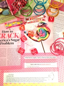 A colorful assortment of candies, including a large rainbow lollipop, Sassy Tarts, Smarties, and Starburst, is displayed on top of a magazine page discussing sugar addiction. Pink, green, and blue plastic spiral toys are placed around the candies. The page contains information about sugar consumption, a quiz on sugar addiction, and a recipe for crack pie.