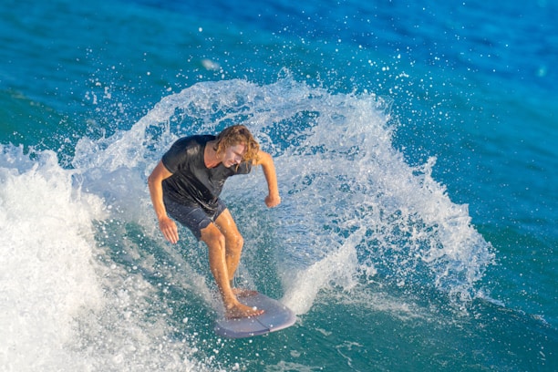 A surfer rides a powerful wave, creating a dramatic splash of water around them. The ocean is vibrant blue, and the person appears focused and skillful, balancing on the surfboard with precision.