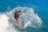 A surfer rides a powerful wave, creating a dramatic splash of water around them. The ocean is vibrant blue, and the person appears focused and skillful, balancing on the surfboard with precision.