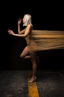A person with white hair is posed in a mid-movement stance, partially wrapped in a sheer, golden fabric. The fabric creates a sense of motion as it trails behind them, contrasting with the dark background. Their stance is energetic, balancing on one foot positioned on a textured floor with a yellow line.