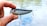 A hand holding a shiny fishing lure with red eyes and two treble hooks. The background shows a blurred view of water, suggesting an outdoor setting.