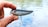 A hand holding a shiny fishing lure with red eyes and two treble hooks. The background shows a blurred view of water, suggesting an outdoor setting.