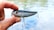 A hand holding a shiny fishing lure with red eyes and two treble hooks. The background shows a blurred view of water, suggesting an outdoor setting.