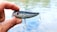 A hand holding a shiny fishing lure with red eyes and two treble hooks. The background shows a blurred view of water, suggesting an outdoor setting.