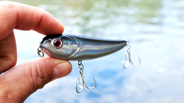 A hand holding a shiny fishing lure with red eyes and two treble hooks. The background shows a blurred view of water, suggesting an outdoor setting.