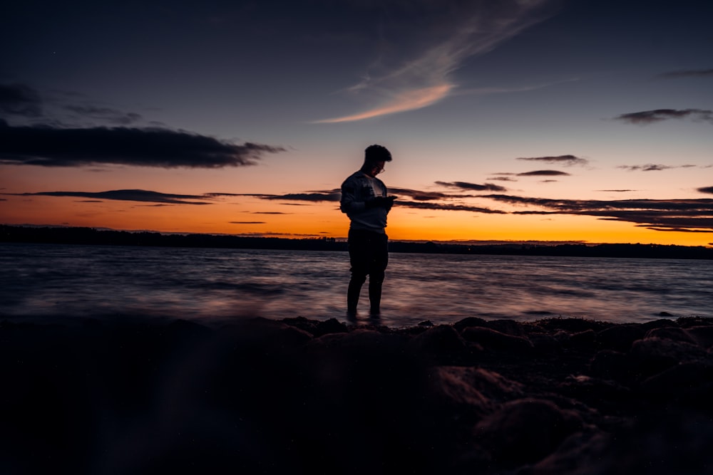 silhouette of woman standing on rocky shore during sunset