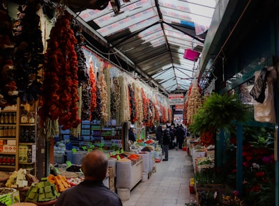 A bustling market aisle filled with various colorful goods, including hanging dried peppers and other produce. The ceiling is made of translucent panels allowing natural light to illuminate the aisles. There are people walking in the corridor, browsing the items displayed on either side.