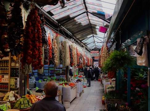A bustling market aisle filled with various colorful goods, including hanging dried peppers and other produce. The ceiling is made of translucent panels allowing natural light to illuminate the aisles. There are people walking in the corridor, browsing the items displayed on either side.