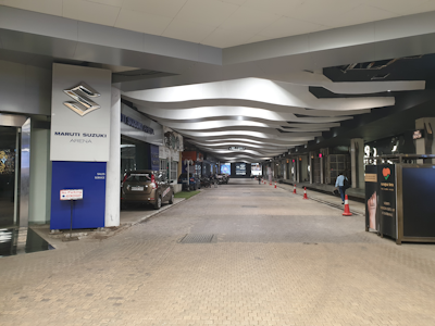 A covered pathway flanked by car showrooms and service centers. The ceiling features wavy, modern design elements. Several vehicles are parked along the side, and orange traffic cones line the edge of the path. The area appears well-lit, with advertising signage on display.