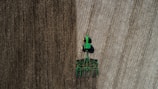 Aerial view of a green tractor tilling a large field with visible rows. The field appears divided by two distinct textures, indicating different stages or types of soil preparation. The tractor and machinery create parallel lines, reflecting the precision of agricultural work.