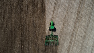 Aerial view of a green tractor tilling a large field with visible rows. The field appears divided by two distinct textures, indicating different stages or types of soil preparation. The tractor and machinery create parallel lines, reflecting the precision of agricultural work.