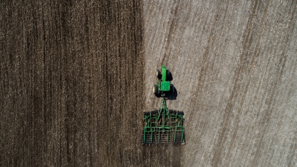 Aerial view of a green tractor tilling a large field with visible rows. The field appears divided by two distinct textures, indicating different stages or types of soil preparation. The tractor and machinery create parallel lines, reflecting the precision of agricultural work.