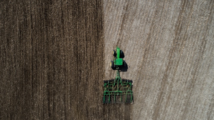 Aerial view of a green tractor tilling a large field with visible rows. The field appears divided by two distinct textures, indicating different stages or types of soil preparation. The tractor and machinery create parallel lines, reflecting the precision of agricultural work.
