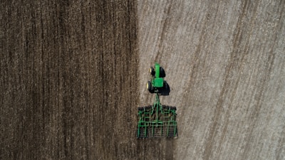 Aerial view of a green tractor tilling a large field with visible rows. The field appears divided by two distinct textures, indicating different stages or types of soil preparation. The tractor and machinery create parallel lines, reflecting the precision of agricultural work.