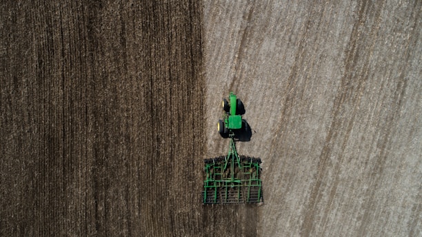 Aerial view of a green tractor tilling a large field with visible rows. The field appears divided by two distinct textures, indicating different stages or types of soil preparation. The tractor and machinery create parallel lines, reflecting the precision of agricultural work.