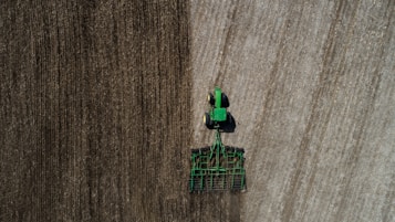 Aerial view of a green tractor tilling a large field with visible rows. The field appears divided by two distinct textures, indicating different stages or types of soil preparation. The tractor and machinery create parallel lines, reflecting the precision of agricultural work.