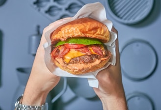 A freshly prepared hamburger is held between two hands. The burger features a toasted bun, a beef patty with melted cheese, crispy bacon, lettuce, and tomato, all neatly wrapped in white paper. The background consists of a blurred gray surface with circular patterns.