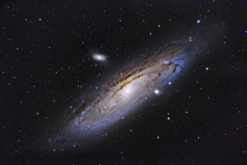 A spiral galaxy is seen against a backdrop of countless stars in deep space. The galaxy has a bright central core surrounded by swirling arms of dust and gas that appear blue and brown. The surrounding stars vary in brightness and color, creating a dynamic and complex celestial scene.
