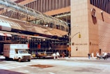 A bustling urban street scene featuring a corner of the World Trade Center with distinctive modern architecture, including a glass-covered pedestrian bridge. People walk along the sidewalk, and vehicles such as a delivery truck and taxi are in the street. The reflection of the buildings emphasizes the structure's height and intricate design.