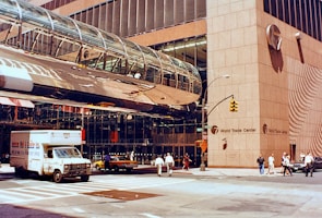 A bustling urban street scene featuring a corner of the World Trade Center with distinctive modern architecture, including a glass-covered pedestrian bridge. People walk along the sidewalk, and vehicles such as a delivery truck and taxi are in the street. The reflection of the buildings emphasizes the structure's height and intricate design.