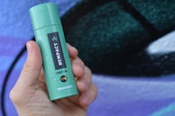 A hand is holding a green cylindrical container labeled 'HEMPACT CBD OIL 10%' with '1000mg Cannabidiol' written on it, against a blurred multicolored background.