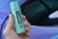 A hand is holding a green cylindrical container labeled 'HEMPACT CBD OIL 10%' with '1000mg Cannabidiol' written on it, against a blurred multicolored background.