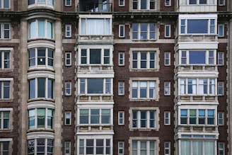 The image features the facade of a multi-story residential building with a classic architectural style. The structure is made of red brick with numerous windows framed by stone or concrete elements. The windows vary in style, with some sections having three or four panels, and others featuring bay windows with rounded shapes. The building has a symmetrical, solid appearance typical of urban architecture from the early to mid-20th century.