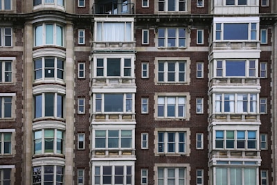 The image features the facade of a multi-story residential building with a classic architectural style. The structure is made of red brick with numerous windows framed by stone or concrete elements. The windows vary in style, with some sections having three or four panels, and others featuring bay windows with rounded shapes. The building has a symmetrical, solid appearance typical of urban architecture from the early to mid-20th century.