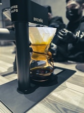 A coffee brewing device labeled 'BREWIE' is situated on a countertop. It features a glass vessel filled with a warm amber liquid and topped with a drip filter holding coffee grounds. In the background, two people wearing black masks and gloves are preparing or observing the process.