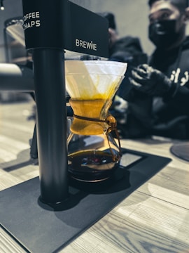 A coffee brewing device labeled 'BREWIE' is situated on a countertop. It features a glass vessel filled with a warm amber liquid and topped with a drip filter holding coffee grounds. In the background, two people wearing black masks and gloves are preparing or observing the process.